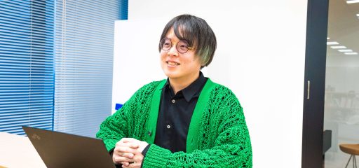 明るくミニマルなデザインのオフィス空間で、鮮やかな緑色のニットカーディガンを着た眼鏡をかけた社員が、ラップトップと共にデスクに座って微笑んでいます。